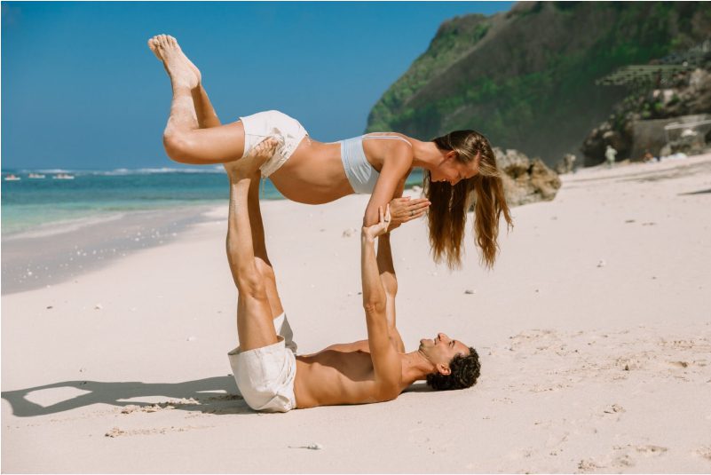 A couple in white shirts and blue jeans doing a partner yoga pose on the sand near the ocean. They are holding each other’s hands and feet and lifting their legs up in the air.