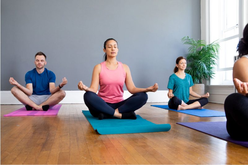 A group of people in various yoga outfits sitting on colorful yoga mats in a bright studio with windows and plants. They are doing a meditation pose with their eyes closed and their hands on their knees.