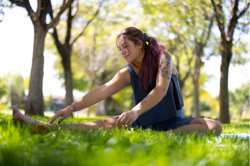 A woman in a blue tank top and black leggings doing a downward-facing dog pose on a green yoga mat in a park with trees and grass in the background.